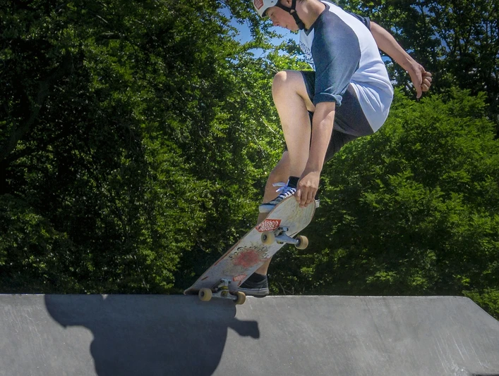 Skatepark Ein Skateboarder mit Helm macht einen Trick auf einer Rampe bei strahlendem Sonnenschein, umgeben von grünen Bäumen.