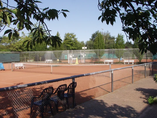 Tennisplatz Tennisplatz mit rotem Sand, umgeben von Bäumen, Zaun im Vordergrund, leere Bänke am Rand.