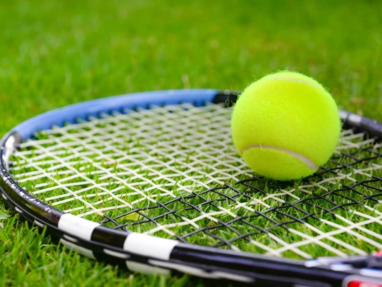 Tennis Ein gelber Tennisball liegt auf einem schwarzen Tennisschläger, der auf grünem Rasen positioniert ist.