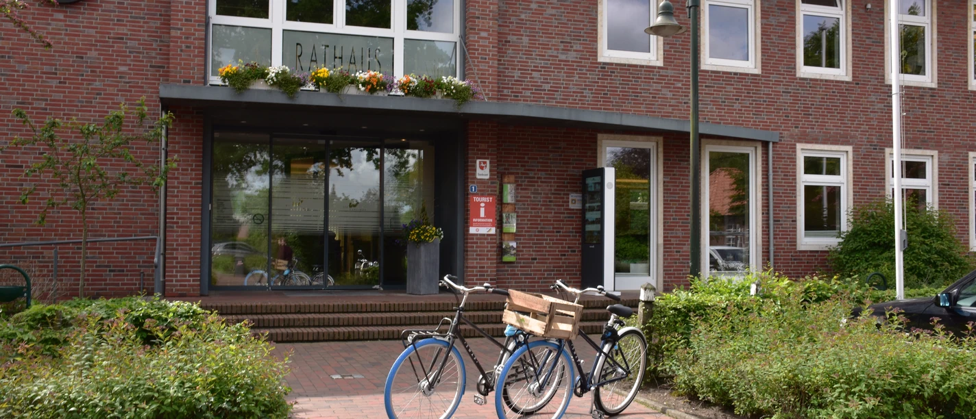 Rathaus Das Bild zeigt ein modernes, zweistöckiges Gebäude aus rotem Backstein mit großen Fenstern. Vor dem Eingang stehen Fahrräder auf einem gepflasterten Weg.