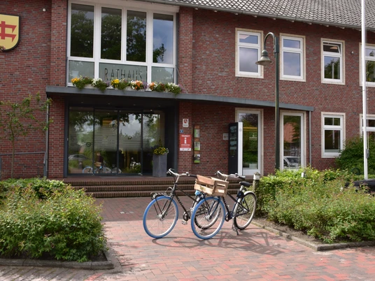 Rathaus Das Bild zeigt ein modernes, zweistöckiges Gebäude aus rotem Backstein mit großen Fenstern. Vor dem Eingang stehen Fahrräder auf einem gepflasterten Weg.