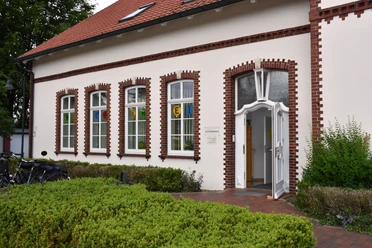 Gemeindebücherei Wiefelstede Das Bild zeigt die Gemeindebücherei Wiefelstede, ein Backsteingebäude mit weißen Fensterrahmen. Vor dem Eingang blühen Sträucher.