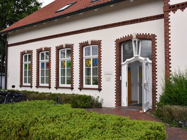 Gemeindebücherei Wiefelstede Das Bild zeigt die Gemeindebücherei Wiefelstede, ein Backsteingebäude mit weißen Fensterrahmen. Vor dem Eingang blühen Sträucher.