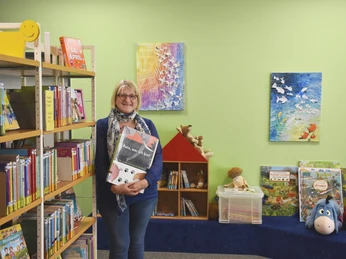 Gemeindebücherei Wiefelstede Eine lächelnde Frau steht vor Bücherregalen in einem gemütlichen Raum mit grüner Wand. Bilder und Spielzeug sind dekorativ angebracht.