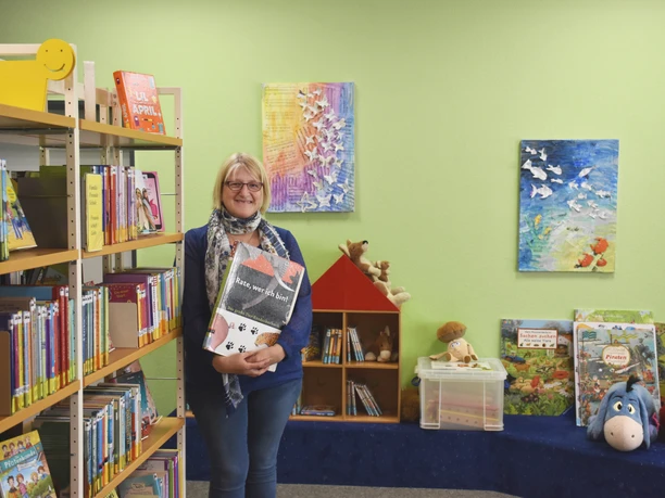Gemeindebücherei Wiefelstede Eine lächelnde Frau steht vor Bücherregalen in einem gemütlichen Raum mit grüner Wand. Bilder und Spielzeug sind dekorativ angebracht.