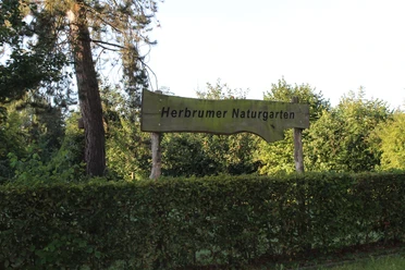 Herbrumer Naturgarten Holzschild mit der Aufschrift „Herbrumer Naturgarten“ vor grünen Bäumen und Hecke im Sonnenlicht