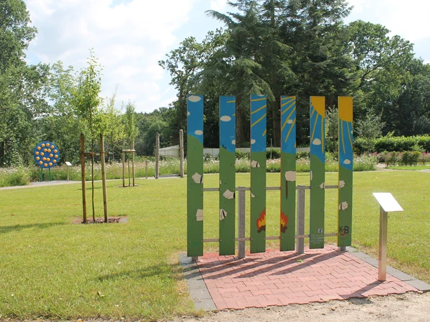 Kolpingwiese Bokeloh Bunte Holzskulptur mit Natur- und Sonnensymbolen auf einer grünen Wiese mit jungen Bäumen.