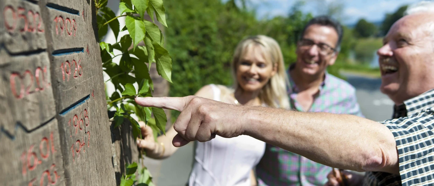 Drei Menschen vor einer geschnitzten Holzstele mit Hochwasserständen