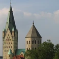 ab_paderborn_dom.jpg Zwei gotische Kirchtürme erheben sich eindrucksvoll gegen einen blauen Himmel, umgeben von Bäumen.