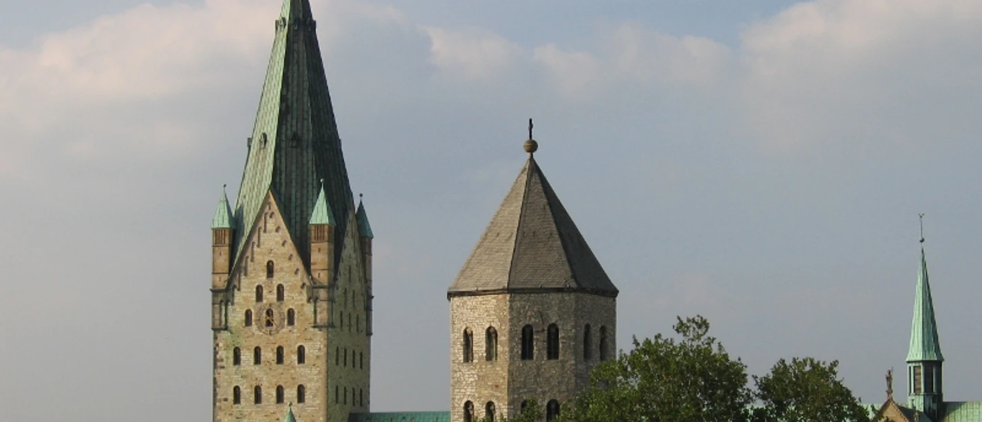 ab_paderborn_dom.jpg Zwei gotische Kirchtürme erheben sich eindrucksvoll gegen einen blauen Himmel, umgeben von Bäumen.