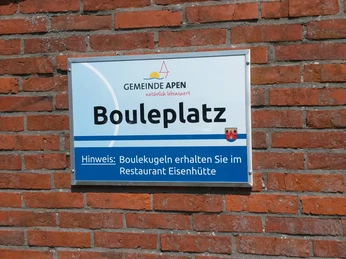 Platzschild Das Bild zeigt ein Schild mit der Aufschrift "Gemeinde Apen - Natürlich lebenswert - Bouleplatz". Unten steht der Hinweis, dass Boulekugeln im Restaurant Eisenhütte erhältlich sind. Das Schild ist an einer roten Backsteinwand angebracht.