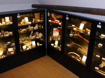 Ausstellung In einem dunklen Raum sind mehrere Vitrinen mit archäologischen Ausstellungsstücken ausgestellt, die beleuchtet sind. Der Raum hat einen hölzernen Fußboden und eine Sichtbalkendecke.