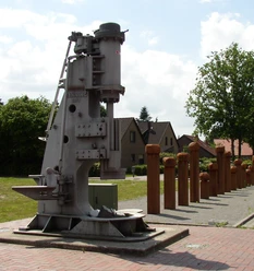 Industriedenkmal_Eisenhüttenplatz Industriedenkmal mit historischem Hammerwerk, eingerahmt von Bäumen und Häusern im Hintergrund.