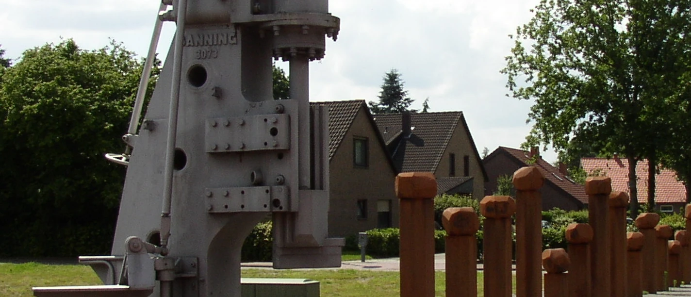 Industriedenkmal_Eisenhüttenplatz Industriedenkmal mit historischem Hammerwerk, eingerahmt von Bäumen und Häusern im Hintergrund.