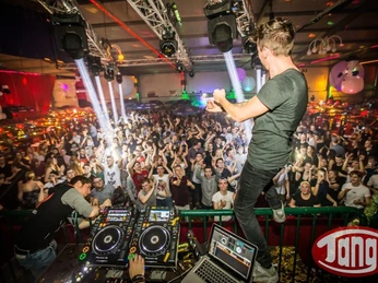 DJ_Disco Tange Die Bildbeschreibung im Alt-Attribut für das Bild "DJ_Disco Tange" könnte folgendermaßen lauten:```htmlEin DJ steht auf einer Bühne, hebt die Hand und blickt auf eine jubelnde Menschenmenge in einem farbenfrohen Club.