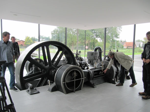 Schauraum Stahlwerk Augustfehn Im Schauraum des Stahlwerks Augustfehn zeigt das Bild eine historische Dampfmaschine mit großen, schwarzen Schwungrädern in einem modernen Ausstellungsraum mit Glaswand. Menschen stehen interessiert um die Maschine.