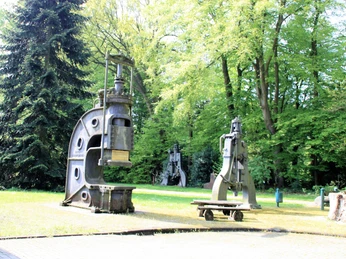 Außengelände Stahlwerk Augustfehn Das Bild zeigt das Außengelände des Stahlwerks Augustfehn. Drei große, historische Metallpressen stehen auf einer gepflegten Rasenfläche, umgeben von hohen Bäumen, die dem Gelände einen parkähnlichen Charakter verleihen. Die Pressen sind imposant und zeugen von der industriellen Vergangenheit des Ortes.