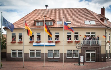 Rathaus_Gemeinde Apen Das Rathaus der Gemeinde Apen, ein zweigeschossiges Gebäude mit rotem Ziegeldach, ist geschmückt mit Blumenkästen und mehreren Flaggenmasten. Vor dem Gebäude stehen Fahnen, inklusive der deutschen und der europäischen Flagge, im leichten Wind flatternd.