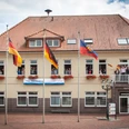 Rathaus_Gemeinde Apen Das Rathaus der Gemeinde Apen, ein zweigeschossiges Gebäude mit rotem Ziegeldach, ist geschmückt mit Blumenkästen und mehreren Flaggenmasten. Vor dem Gebäude stehen Fahnen, inklusive der deutschen und der europäischen Flagge, im leichten Wind flatternd.