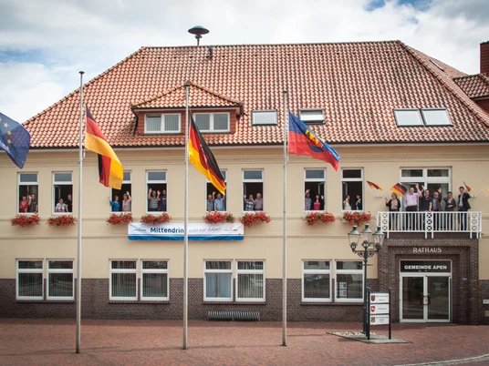 Rathaus_Gemeinde Apen Das Rathaus der Gemeinde Apen, ein zweigeschossiges Gebäude mit rotem Ziegeldach, ist geschmückt mit Blumenkästen und mehreren Flaggenmasten. Vor dem Gebäude stehen Fahnen, inklusive der deutschen und der europäischen Flagge, im leichten Wind flatternd.