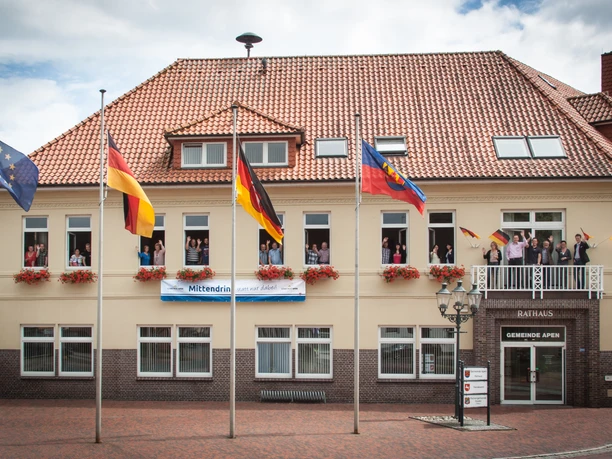 Rathaus_Gemeinde Apen Das Rathaus der Gemeinde Apen, ein zweigeschossiges Gebäude mit rotem Ziegeldach, ist geschmückt mit Blumenkästen und mehreren Flaggenmasten. Vor dem Gebäude stehen Fahnen, inklusive der deutschen und der europäischen Flagge, im leichten Wind flatternd.