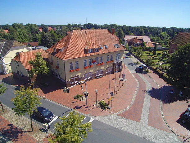 Luftaufnahme_Rathaus_Gemeinde Apen Die Luftaufnahme zeigt das beige Rathaus der Gemeinde Apen mit roten Dachziegeln, umgeben von Bäumen.
