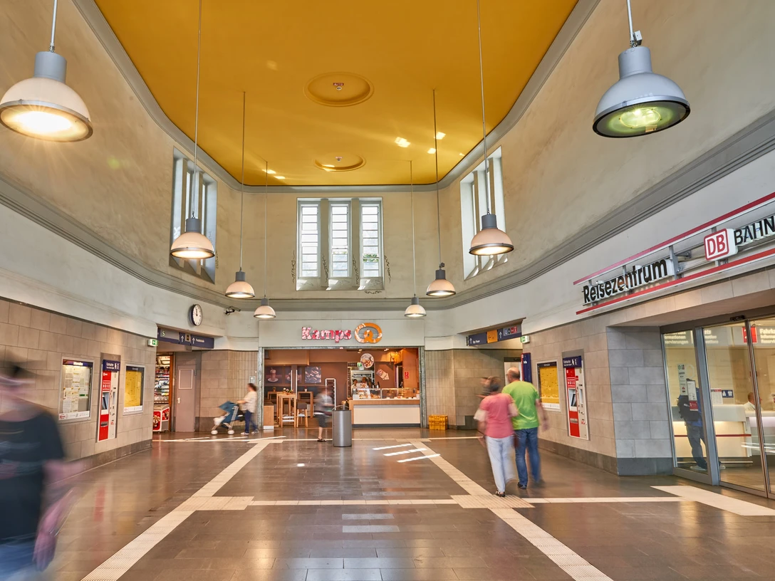 Hauptbahnhof, Mülheim an der Ruhr Großzügige Eingangshalle mit hohen Decken, leuchtend gelber Wandfarbe und Beleuchtung, rechts ein Schalter der Deutschen Bahn.