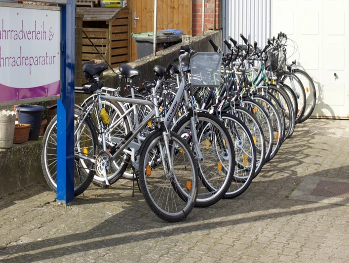 Auswahl der Fahrräder Eine lange Reihe von weißen Fahrrädern steht schräg nebeneinander auf einem gepflasterten Hof. Links ist ein Schild mit der Aufschrift "Fahrradverleih & Fahrradreparatur" sichtbar.