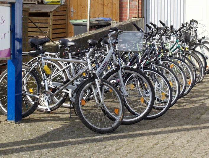 Auswahl der Fahrräder Eine lange Reihe von weißen Fahrrädern steht schräg nebeneinander auf einem gepflasterten Hof. Links ist ein Schild mit der Aufschrift "Fahrradverleih & Fahrradreparatur" sichtbar.