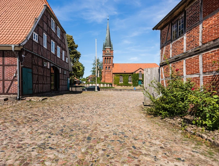 Hippolit Kirche Amelinghausen Das Bild zeigt einen gepflasterten Weg, flankiert von historischen Fachwerkhäusern, der zur beeindruckenden Hippolit Kirche in Amelinghausen führt. Der Kirchturm erhebt sich majestätisch in den klaren blauen Himmel.