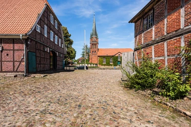 Hippolit Kirche Amelinghausen Das Bild zeigt einen gepflasterten Weg, flankiert von historischen Fachwerkhäusern, der zur beeindruckenden Hippolit Kirche in Amelinghausen führt. Der Kirchturm erhebt sich majestätisch in den klaren blauen Himmel.