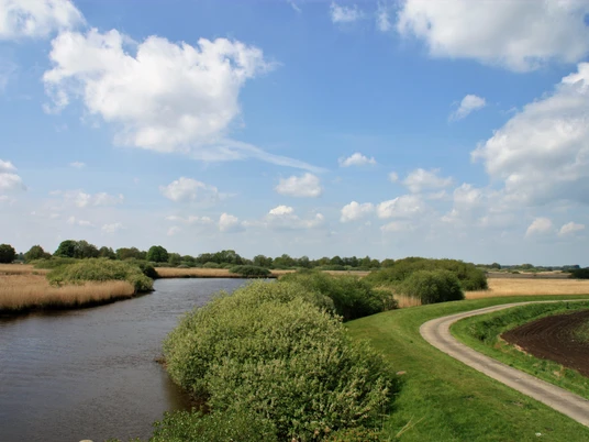 Ausblick_Landschaftsfenster_Tange Sanft geschwungene Flusslandschaft unter blauem Himmel mit weißen Wolken, umgeben von grünen Wiesen und Bäumen.