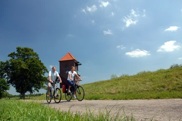 Radlerpaar_Landschaftsfenster_Wasser Ein Radlerpaar fährt bei sonnigem Wetter einen ruhigen Weg entlang, flankiert von grünen Wiesen und einem Baum. Im Hintergrund ist ein gemauerter Turm mit einem roten Dach zu sehen, während leichte Wolken den blauen Himmel zieren.