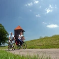 Radlerpaar_Landschaftsfenster_Wasser Ein Radlerpaar fährt bei sonnigem Wetter einen ruhigen Weg entlang, flankiert von grünen Wiesen und einem Baum. Im Hintergrund ist ein gemauerter Turm mit einem roten Dach zu sehen, während leichte Wolken den blauen Himmel zieren.