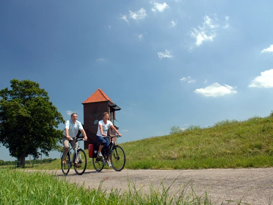 Radlerpaar_Landschaftsfenster_Wasser Ein Radlerpaar fährt bei sonnigem Wetter einen ruhigen Weg entlang, flankiert von grünen Wiesen und einem Baum. Im Hintergrund ist ein gemauerter Turm mit einem roten Dach zu sehen, während leichte Wolken den blauen Himmel zieren.