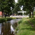 Augustfehn Kanal mit Treidelpfad Ein idyllischer Kanal fließt unter einer modernen Fußgängerbrücke, begleitet von einem erholsamen Treidelpfad und gesäumt von Bäumen. Der Augustfehn Kanal bietet eine malerische Kulisse für Spaziergänger und Naturliebhaber.
