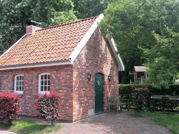 Backhaus an der Hengstforder Mühle Ein Backhaus aus rotem Ziegelstein mit Satteldach, umgeben von grünen Bäumen und roten Sträuchern.
