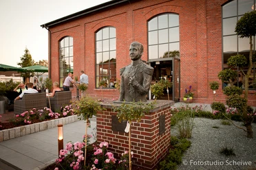 Bronzebüste Paul Friedrich August Die Bronzebüste von Paul Friedrich August steht prominent vor einem rot verklinkerten Gebäude. Der Kopf der Büste ist detailreich gestaltet und fängt den Ausdruck des berühmten Figuren gut ein. Umgeben von gepflegten Blumenbeeten und gemütlichen Sitzgelegenheiten, wirkt der Ort einladend und harmonisch.