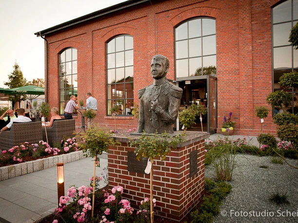 Bronzebüste Paul Friedrich August Die Bronzebüste von Paul Friedrich August steht prominent vor einem rot verklinkerten Gebäude. Der Kopf der Büste ist detailreich gestaltet und fängt den Ausdruck des berühmten Figuren gut ein. Umgeben von gepflegten Blumenbeeten und gemütlichen Sitzgelegenheiten, wirkt der Ort einladend und harmonisch.