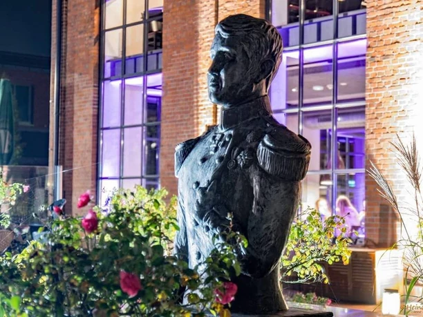 Büste Großherzog Das Bild zeigt eine Bronze-Büste eines Großherzogs vor einer modernen Gebäude-Fassade bei Nacht, umgeben von Pflanzen und Rosen.