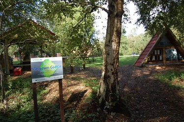 np-moor_weg-zum-glueck_3 Holzschild mit Aufschrift „Start Weg zum Glück“ vor Bäumen und einer kleinen Holzhütte im Grünen