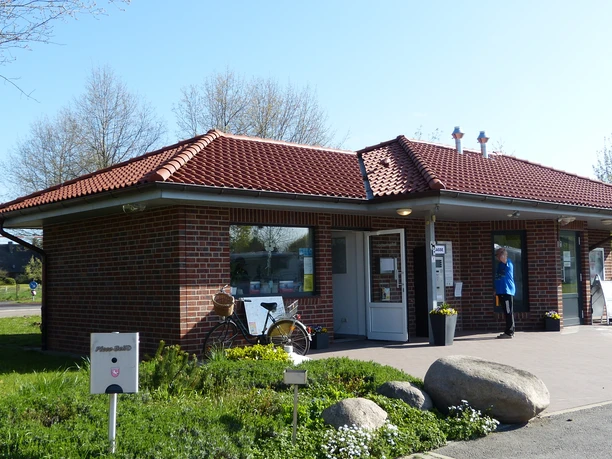 Das Servicegebäude auf dem Wohnmobilstellplatz besitzt ein rotes Ziegeldach und Backsteinwände, flankiert von Fenstern und einer Tür. Ein Fahrrad steht an der Wand, umgeben von grüner Vegetation und einem klaren blauen Himmel, das eine einladende Atmosphäre verbreitet.
