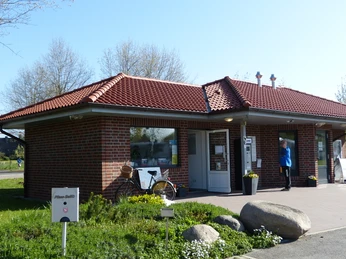 Servicegebäude Wohnmobilstellpatz Das Servicegebäude auf dem Wohnmobilstellplatz besitzt ein rotes Ziegeldach und Backsteinwände, flankiert von Fenstern und einer Tür. Ein Fahrrad steht an der Wand, umgeben von grüner Vegetation und einem klaren blauen Himmel, das eine einladende Atmosphäre verbreitet.