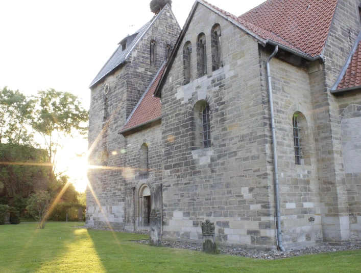 Die Sigwardskirche, ein historisches Steingebäude, wird im Abendlicht von der golden untergehenden Sonne angestrahlt, was die Struktur und die Details der gotischen Architektur betont. Links lehnt sich ein Schatten entlang des Rasens, der das friedliche Ambiente untermalt.
