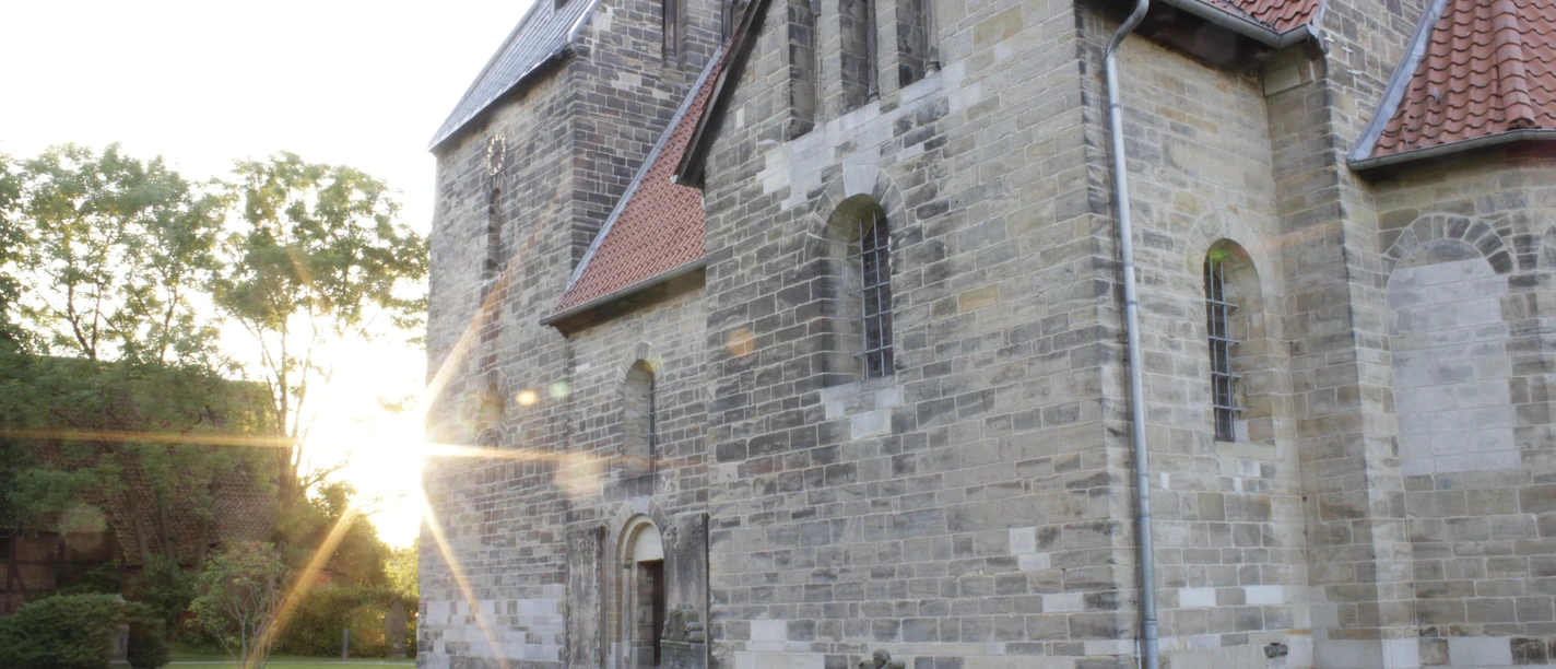 Die Sigwardskirche, ein historisches Steingebäude, wird im Abendlicht von der golden untergehenden Sonne angestrahlt, was die Struktur und die Details der gotischen Architektur betont. Links lehnt sich ein Schatten entlang des Rasens, der das friedliche Ambiente untermalt.