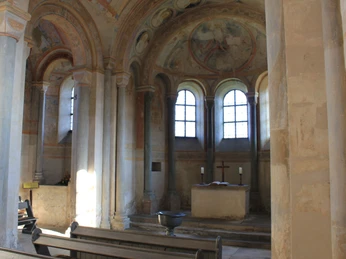 Der Altarraum der Sigwardskirche zeigt eine schlichte, romanische Architektur mit einem zentralen Altar. Große Bogenfenster lassen sanftes Licht in den Raum strömen, wobei Fresken mit biblischen Szenen die Wände zieren und eine ruhige, sakrale Atmosphäre schaffen. Der Raum ist von Säulen umgeben, die den Altar eindrucksvoll rahmen.