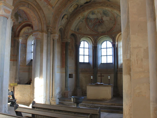 Altarraum Sigwardskirche Der Altarraum der Sigwardskirche zeigt eine schlichte, romanische Architektur mit einem zentralen Altar. Große Bogenfenster lassen sanftes Licht in den Raum strömen, wobei Fresken mit biblischen Szenen die Wände zieren und eine ruhige, sakrale Atmosphäre schaffen. Der Raum ist von Säulen umgeben, die den Altar eindrucksvoll rahmen.