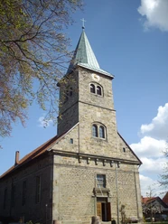 Steinernes Kirchenschiff mit Kirchturm, schiefer Spitze und Uhr unter blauem Himmel mit Wolken.