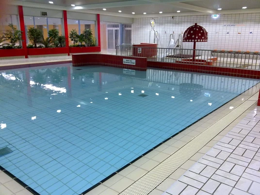 Wunstorf Elements Nichtschwimmerbecken Das Bild zeigt ein modernes Hallenbad mit einem nicht zu tiefen Beckenbereich. Die Fliesen haben eine helle, glänzende Oberfläche und sind in neutralen Tönen gehalten, was eine saubere und einladende Atmosphäre schafft. Pflanzen und Sitzgelegenheiten ergänzen das freundliche Ambiente.