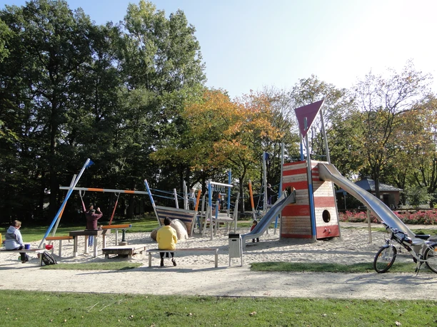 Verschiedene bunte und moderne Spielgeräte auf einem Sandspielplatz, umgeben von Grünflächen.